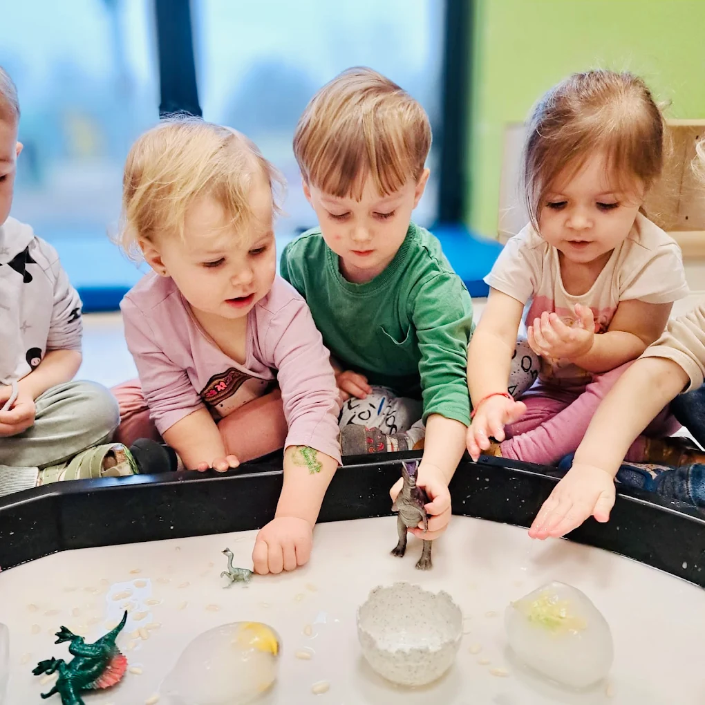Troje dzieci bawi się w tacy sensorycznej z figurkami dinozaurów i zamrożonymi jajkami